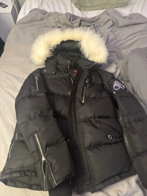 Svart pufferjacka från Moose Knuckles - Svart pufferjacka från Moose Knuckles med dunfyllning och stor huva med vit päls. Jackan har flera dragkedjor, ribbade muddar och snygga detaljer på ärmen. Perfekt för kalla dagar med sin tjocka och quiltade design.litet hål i fickan men inget större de blir ba som större fickor. Pris kan diskuteras