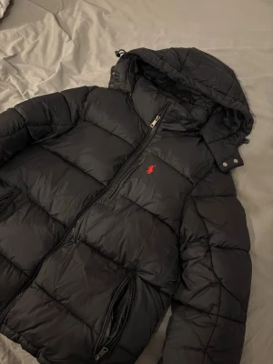 Svart dunjacka från Polo Ralph Lauren - Snygg svart dunjacka från Polo Ralph Lauren med röd broderad logga på bröstet. Jackan har huva, dragkedja framtill och två sidofickor med dragkedja. Perfekt för kalla vinterdagar och ger en stilren look.