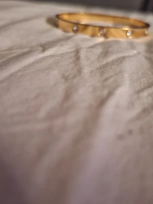 Guldigt stelt armband med detaljer - Snyggt stelt armband i guldfärgad metall med små runda detaljer runt om. Perfekt accessoar för att lyfta din outfit med en stilren och modern touch. Armbandet har en enkel och elegant design som passar till många olika stilar.