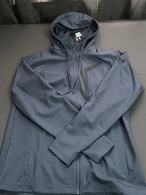 Mörkgrå hoodie från Under Armour - Snygg mörkgrå hoodie från Under Armour med dragkedja framtill och justerbar huva. Tillverkad i ett mjukt och stretchigt material som känns sportigt. Har en diskret logga på ärmen och en cool detalj på bröstet. Perfekt för en avslappnad och sportig stil.