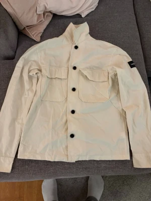 Beige overshirt från Calvin Klein - Stilren beige overshirt från Calvin Klein med raka linjer och två stora bröstfickor. Skjortan har svarta knappar framtill och en diskret logga på ärmen. Tillverkad i ett lätt och slitstarkt material, perfekt för lager på lager.