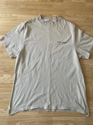 Beige t-shirt från Samsøe Samsøe - Snygg beige t-shirt från Samsøe Samsøe med minimalistiskt tryck på bröstet. Klassisk passform med rund hals och korta ärmar. Tillverkad i mjuk bomull som känns skön mot huden. Perfekt basplagg till garderoben. Passar mig som är 183 fast sitter lite oversize. 