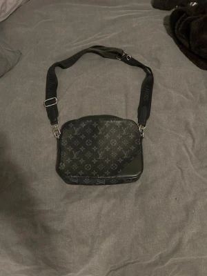 Svart axelväska Louis Vuitton - Snygg svart axelväska från Louis Vuitton med klassiskt monogrammönster och justerbar axelrem. Väskan har en rektangulär form och detaljer i läder. Perfekt för dig som vill ha en stilren och lyxig accessoar.