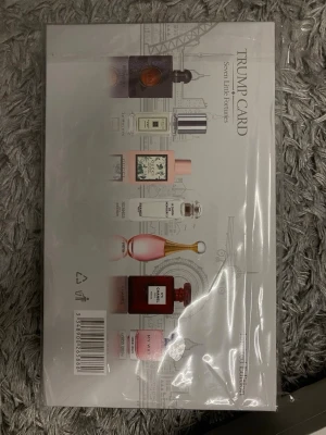 Trump Card Seven Little Fortunes parfymset - Snyggt parfymset från Trump Card med sju olika dofter i små flaskor. Förpackningen är vit med gulddetaljer och har en elegant stadssilhuett på framsidan. Perfekt för dig som vill testa flera olika parfymer och hitta din favorit.