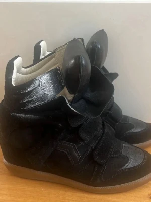 Svarta Isabel Marant wedge sneakers - Svarta wedge sneakers från Isabel Marant med högt skaft och kardborreband. Skorna har en blandning av mocka och läder, samt en cool design med vadderad plös och gummisula. Perfekta för dig som vill sticka ut med din stil.