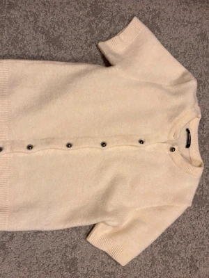 Stickad kofta - Kortärmad krämvit kofta från Gina Tricot med rund halsringning och silvriga knappar framtill. Mjuk stickad känsla och ribbade kanter vid ärmslut och nederkant. Perfekt för lager-på-lager och enkel att matcha med olika outfits.