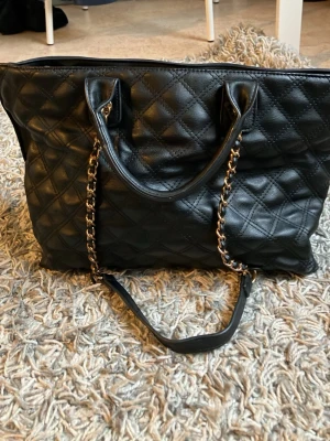 Svart handväska med guldkedja från Don Donna - Snygg svart handväska från Don Donna i slätt konstläder. Väskan har både handtag och en axelrem med guldkedja, samt dragkedja upptill. Rymlig insida med svart foder och flera fack för småsaker. Perfekt för dig som vill ha plats för allt du behöver.