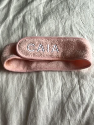 Rosa hårband från CAIA - Säljer ett mjukt och stretchigt rosa hårband från CAIA med vit broderad logga framtill. Perfekt för att hålla håret borta vid sminkning eller hudvård. Kardborreknäppning gör det enkelt att justera storleken. Materialet är frottéliknande och bekvämt mot huden.