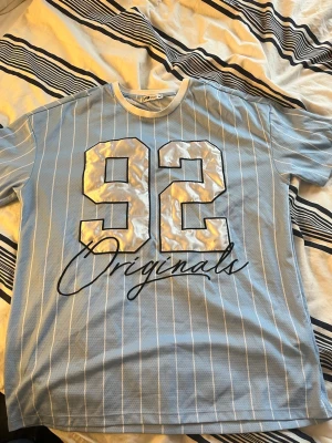 Blå randig t-shirt Originals 92 - Snygg ljusblå t-shirt med vita ränder och stort tryck med siffran 92 och texten 'Originals' på framsidan. Klassisk rund halsringning och korta ärmar. Perfekt för en avslappnad streetstyle-look. Lite åt de oversized hållet. 100% polyester.