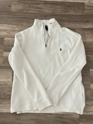 Vit half-zip tröja från Polo Ralph Lauren - Klassisk vit långärmad tröja från Polo Ralph Lauren med half-zip och broderad logga på bröstet. Tröjan har ribbade muddar och är tillverkad i mjukt material som passar perfekt till en avslappnad men stilren look.