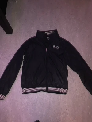 Svart EA7 vindjacka med randiga detaljer - Svart vindjacka från EA7 Emporio Armani med dragkedja framtill och vita logotypdetaljer på bröstet. Jackan har randiga muddar i svart, vitt och rosa vid ärmslut och midja samt ståkrage med randigt foder. Perfekt för en sportig och stilren look.