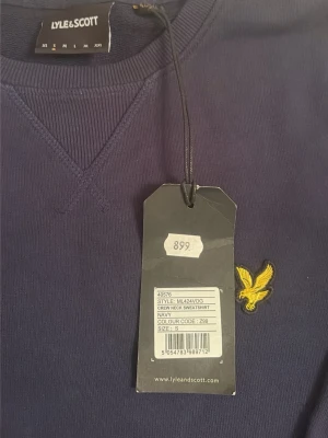 Marinblå sweatshirt från Lyle & Scott - Klassisk marinblå sweatshirt från Lyle & Scott med rund halsringning och det ikoniska gula örnlogot på bröstet. Tröjan har ribbade muddar vid ärmslut och nederkant samt en diskret V-detalj vid halsen. Perfekt för en stilren och avslappnad look.