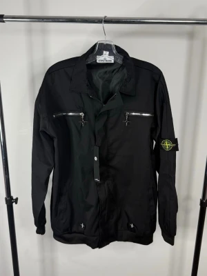  Stone Island sommar jacka - Svart jacka från Stone Island med klassisk krage, två bröstfickor med dragkedjor och ikonisk logopatch på ärmen. Tillverkad i ett slitstarkt material med långa ärmar och justerbar nederkant. Perfekt för dig som gillar streetwear och stilrena detaljer.