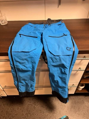 Blå täckbyxor från Norrøna - Snygga blå täckbyxor från Norrøna med svarta detaljer och dragkedjor längs benen. Byxorna har stora fickor med dragkedja framtill och förstärkta benslut. Perfekta för vinterns äventyr i backen eller på fjället.