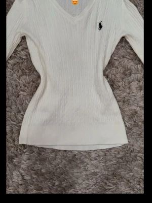 Vit kabelstickad tröja Ralph Lauren - Snygg vit kabelstickad tröja från Ralph Lauren med V-ringning och svart broderad logga på bröstet. Tröjan har lång ärm och smal passform, perfekt till jeans eller kjol. Klassisk och stilren design som aldrig går ur tiden.