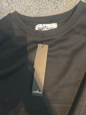 Svart långärmad tröja Stone Island - Svart långärmad tröja från Stone Island med klassisk rund halsringning och ikonisk logopatch på ärmen. Tröjan är tillverkad i mjukt material och har en stilren design som passar perfekt till streetwear. Taggen sitter kvar.