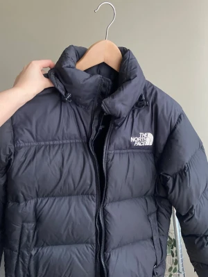 Svart dunjacka från The North Face - svart dunjacka från The North Face med hög krage, dragkedja och justerbar huva. Jackan har en puffig design och logga på bröstet. Använd i en säsong och är i bra skick, köpt för 3800 och den är svår att få tag på i butik! 