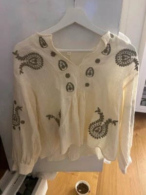 Beige blus med paisleymönster - Luftig beige blus med gröna  paisleyinspirerade broderier och v-ringning. Köpt på marknad utomlands så vet inte vart den är ifrån. Endast använd 1 gång säljer då jag inte använde den. Pris kan diskuteras vid snabb affär. Skriv till mig vid minsta fråga.