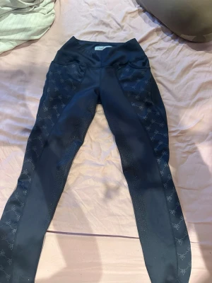 Svarta ridleggings Cavalli Puri - Snygga svarta ridleggings från Cavalli Puri med hög midja och silikonprint längs benen för extra grepp. Diskreta mönsterdetaljer på sidorna och stretchigt material som ger skön passform. Perfekta för dig som vill ha både stil och funktion i stallet.