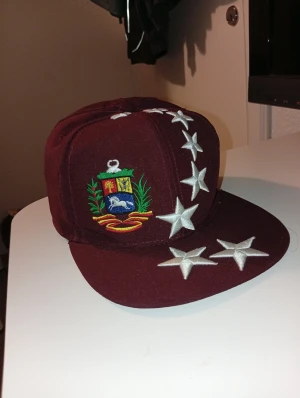 Vinröd keps med broderade stjärnor - Säljer en vinröd snapback-keps med broderade vita stjärnor över skärmen och framsidan. På sidan finns ett färgglatt broderat emblem med sköld, häst och växter. Kepsen har platt skärm och justerbar baksida. Perfekt för dig som vill sticka ut med unik stil.