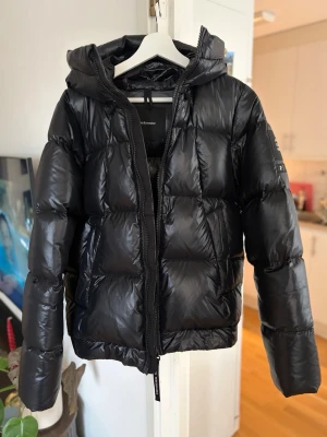 Svart pufferjacka från Peak Performance - Snygg svart pufferjacka från Peak Performance med huva och dragkedja framtill. Jackan har en quiltad design med stora paneler och logotyp på ärmen. Perfekt för kalla vinterdagar och riktigt varm tack vare sitt vadderade material.