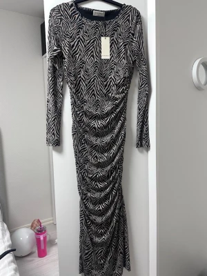 Zebramönstrad långklänning - ny - Säljer en långärmad långklänning  med zebramönster i svart och beige. Klänningen har åtsittande passform och rynkade detaljer längs sidorna för en snygg siluett. Perfekt för dig som vill sticka ut med ett djurmönster!
