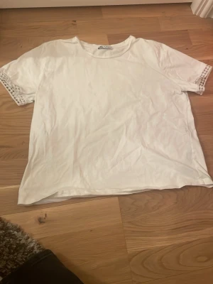 Zara T-shirt  - Säljer en enkel vit t-shirt med korta ärmar och snygga spetsdetaljer längst ut på ärmarna. Perfekt till jeans eller kjol för en clean och fräsch look. Materialet känns mjukt och skönt mot huden.