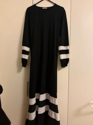 Svart långklänning med vita ränder - Säljer en svart långklänning från Adidas med vita ränder på ärmar och nederkant. Klänningen har lång ärm, rund halsringning och ett avtagbart bälte i midjan för en snygg siluett. Perfekt för dig som gillar sportig och stilren design.