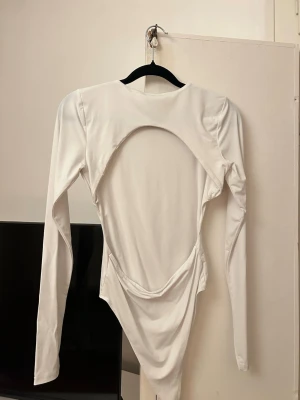 Vit långärmad body från Zara - Snygg vit body från Zara med lång ärm och rund halsringning. Baksidan har en stor, öppen cut-out som ger en trendig look. Tillverkad i ett stretchigt och mjukt material som sitter tight mot kroppen. Perfekt för att skapa en stilren och modern outfit.