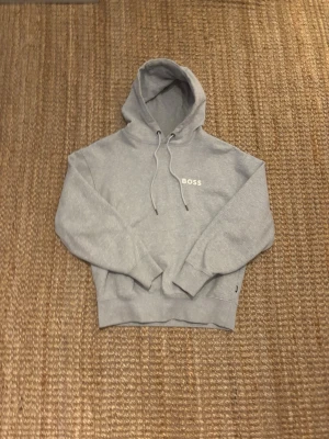 Grå hoodie från BOSS - Snygg grå hoodie från BOSS med klassisk huva och dragsnören. Tröjan har en diskret BOSS-logga på bröstet och en stor magficka framtill. Tillverkad i mjukt material som känns skönt mot huden. Perfekt för en avslappnad och stilren look. 
