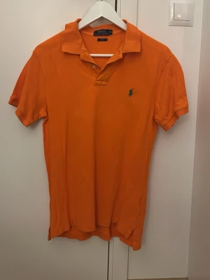 Orange pikétröja från Polo Ralph Lauren - Klassisk orange pikétröja från Polo Ralph Lauren med krage, korta ärmar och den ikoniska gröna logotypen broderad på bröstet. Tillverkad i mjuk bomull och har knappar vid halsen. Perfekt för en avslappnad och stilren look.