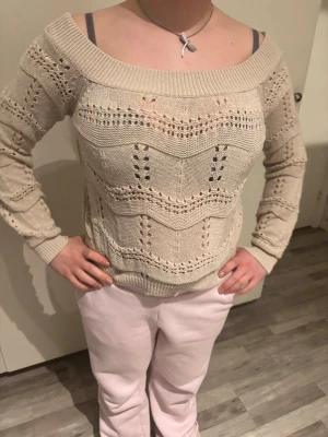 Beige off shoulder stickad tröja - Säljer en beige stickad tröja med bred off shoulder-ringning och hålmönster. Tröjan har långa ärmar, ribbade muddar och ett snyggt vågigt mönster. Perfekt för dig som gillar en avslappnad men trendig stil.