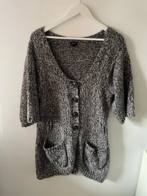 Gråmelerad stickad kofta från Lindex - Säljer en gråmelerad stickad kofta från Lindex med knappar framtill, två stora fickor och halvlånga ärmar samt bälteshällor. Koftan har en djup V-ringning och är gjord i mjukt garn.