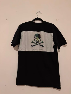 Svart t-shirt från Philipp Plein - Cool svart t-shirt från Philipp Plein med en grå panel på ryggen och en färgglad döskalle- och korslagda ben-tryck. Klassisk passform med rund hals och korta ärmar. Märkeslapp på framsidan. Perfekt för dig som gillar streetwear.