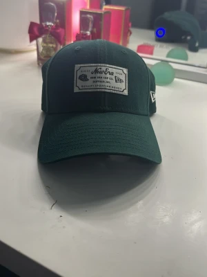 Mörkgrön keps från New Era - Snygg mörkgrön keps från New Era med böjd skärm och justerbar rem bak. Kepsen har en vit patch framtill med logga och text samt klassisk passform. Tillverkad i slitstarkt tyg, perfekt för en casual streetwear-look.
