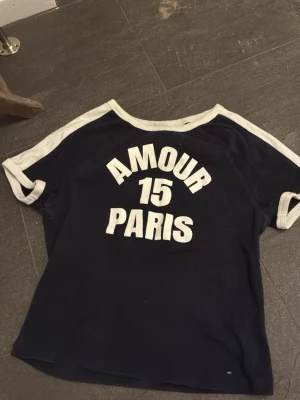 Blå t-shirt med text AMOUR PARIS - Mörkblå t-shirt med vita kontrastkanter och trycket 'AMOUR 15 PARIS' på framsidan. T-shirten har korta ärmar och en avslappnad passform. Perfekt för dig som gillar sportig och trendig stil.