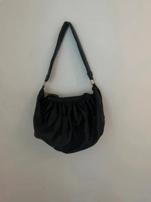 Svart axelväska  - Stilren svart axelväska med mjuk, rynkad design och bred axelrem. 