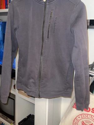 Sail Racing - Snygg mörkgrå hoodie från Sail Racing med dragkedja framtill och en bröstficka med zip. Hoodien har en klassisk huva och diskret logga på bröstet. Tillverkad i mjukt bomullsmaterial, perfekt för chill dagar eller när du vill ha en clean look.