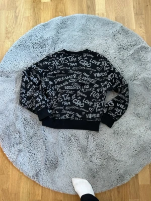 Svart sweatshirt med vit textprint - Cool svart sweatshirt med vit graffiti-inspirerad textprint över hela tröjan. Tröjan har rund hals, långa ärmar och ribbade muddar vid ärmslut och nederkant. Perfekt för en avslappnad streetstyle-look.