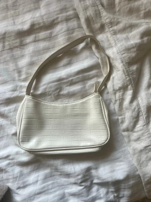Vit axelremsväska i croc-mönster - Säljer en stilren vit handväska med axelrem och croc-embossad yta. Väskan har en enkel, rektangulär form och stängs med dragkedja upptill. 
