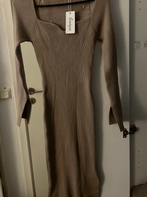 Beige ribbad långklänning Georgia - Snygg beige ribbad långklänning från Georgia med markerad byst och lång ärm. Klänningen har en kroppsnära passform.