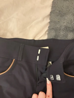 Mörkblå ridbyxor med silikon - Snygga mörkblå ridbyxor med hög midja och silikongrepp längs insidan av benen. Byxorna har två knappar i midjan, bälteshällor och kontrastfärgade detaljer vid fickorna. Perfekta för dig som gillar ridning och vill ha bra grepp i sadeln.
