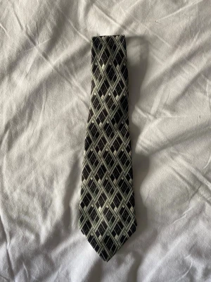 Vintage slips från Carven Paris - Snygg slips från Carven Paris i 100% siden med geometriskt rutmönster i svart, grått och ljusgrönt. Tillverkad i Frankrike och har en klassisk form som passar till skjorta och kavaj. Perfekt accessoar för att lyfta din outfit.