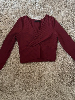 Vinröd omlottopp från SHEIN - Trendig vinröd omlottopp från SHEIN med ribbad struktur och lång ärm. Toppen har en croppad passform och djup v-ringning som ger en snygg siluett. Perfekt att matcha med höga jeans eller kjol för en cool look.