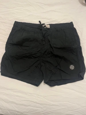 Svarta shorts från Stone Island - Svarta badshorts från Stone Island med elastisk midja och snörning. Klassisk logopatch på benet och praktiska fickor fram. Tillverkade i lätt syntetmaterial med meshfoder, perfekta för varma dagar. Säljer dessa badshorts för att del ej kommer till användning så ofta! Skicket är bra😁🤩