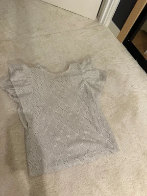 Vit blus med volangärmar och mönster - Söt vit blus med volangdetaljer på ärmarna och ett diskret rutmönster i tyget. Blusen har rund halsringning och är tillverkad i ett lätt material som känns luftigt. Perfekt för dig som gillar feminina detaljer och stilrena plagg.