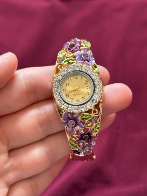 Blommig guldfärgad klocka med stenar - Unik guldfärgad klocka med lila och gröna blommor i emalj och gnistrande stenar runt urtavlan. Klockan har ett dekorativt armband med detaljerade blommor och blad, och en klassisk rund urtavla med guldfärgade visare. Perfekt för dig som gillar färgglada accessoarer.