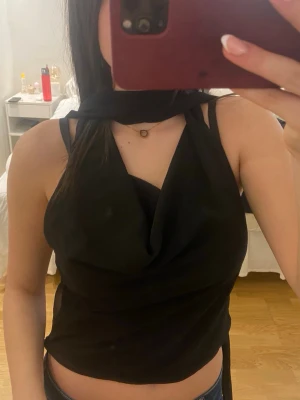 Svart omlott halterneck topp - Snygg svart halterneck topp med omlott-detalj och draperad framsida. Toppen är ärmlös och har en croppad passform som knyts i nacken. Perfekt för att skapa en trendig och stilren look.