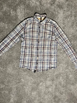 Rutig skjorta från Nudie jeans.co - Rutig skjorta från Nudie jeans Co i blått, beige och orange. Skjortan har klassisk krage, långa ärmar och knäppning framtill. Perfekt för en avslappnad och stilren look.