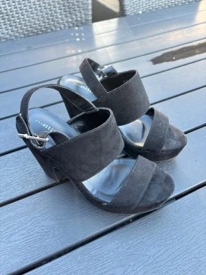 Svarta platåpumps med bred klack - Säljer ett par svarta pumps med bred klack och platåsula. Skorna har två breda remmar över foten och justerbart spänne vid ankeln. Materialet är mockaimitation som ger en snygg och trendig look. Perfekta för dig som vill ha både höjd och stabilitet. Använda ca 2 gånger